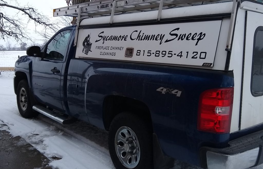 Blue pickup truck with a white topper and ladder rack. The white topper is lettered with "Sycamore Chimney Sweep 815-895-4120" Parked in a snow covered driveway