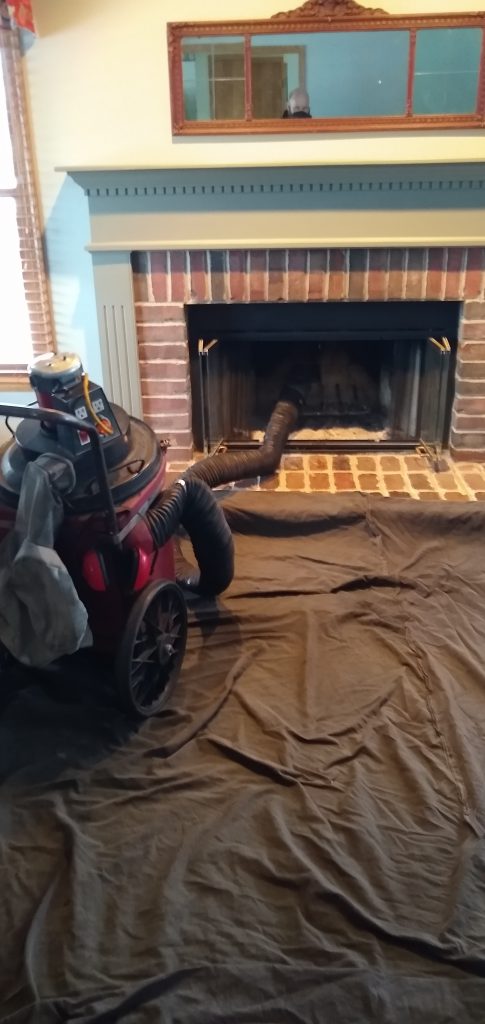 Shopvac on floor. Hose from vaccuum inside a fireplace with brick surround.